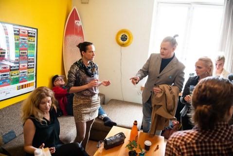 people discussing a topic around a table, some seated, some standing, with a surfboard behind them