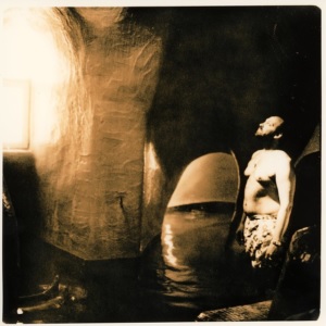 Photo ocre et noir avec un joueur, torse-nu, dans ce qui semble être une grotte naturelle. L'eau lui arrive aux genoux et il regarde la lumière qui entre par une ouverture.