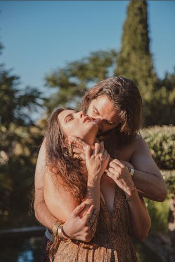 Photo d'un couple s'enlaçant toridement en extérieur. L'un, torse nu, fait un baiser dans le cou à l'autre, qui ferme les yeux en extase.