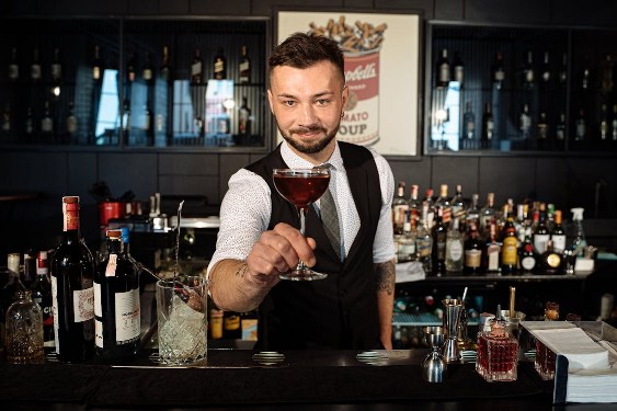 barman présentant un cocktail, devant une collection de bouteilles d'alcool évoquant les JdR dans la bibliothèque d'un rôliste