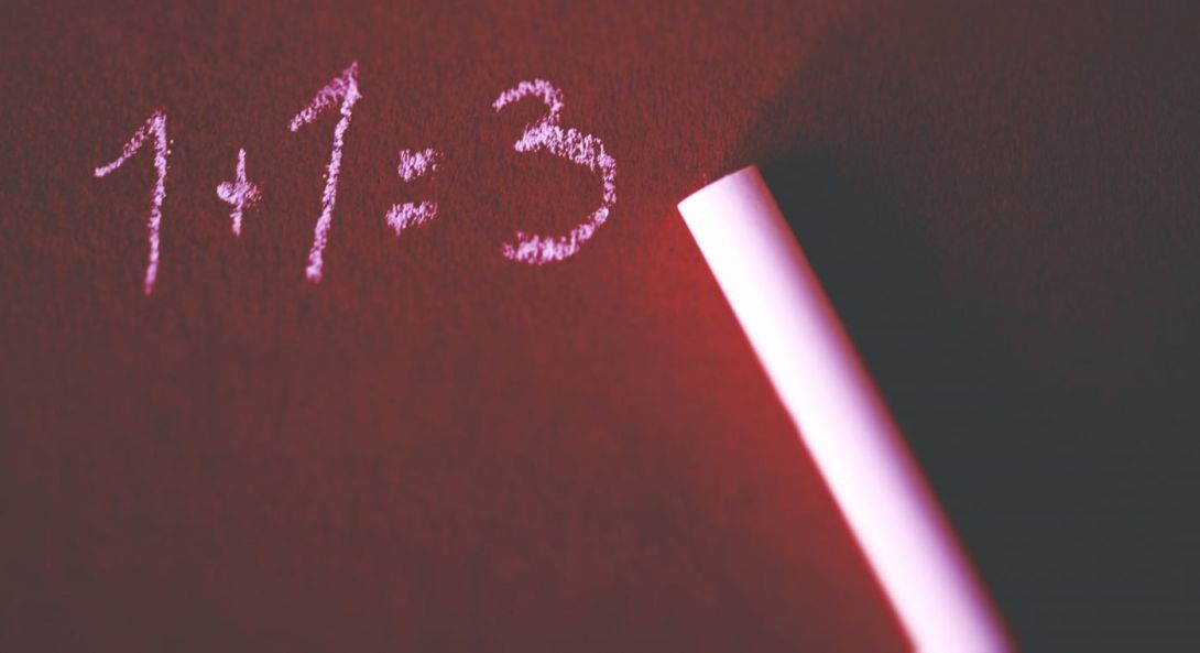 Un tableau en ardoise illuminé par une lumière rougeâtre. À côté du bâton de craie posé sur le tableau, on voit l'addition 1+1=3 écrite avec cette craie.