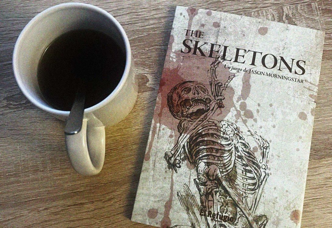 photo d'un mug de café, posé sur une table en bois, à côté du (petit) livre de JdR "The Skeletons", un jeu de Jason Morningstar